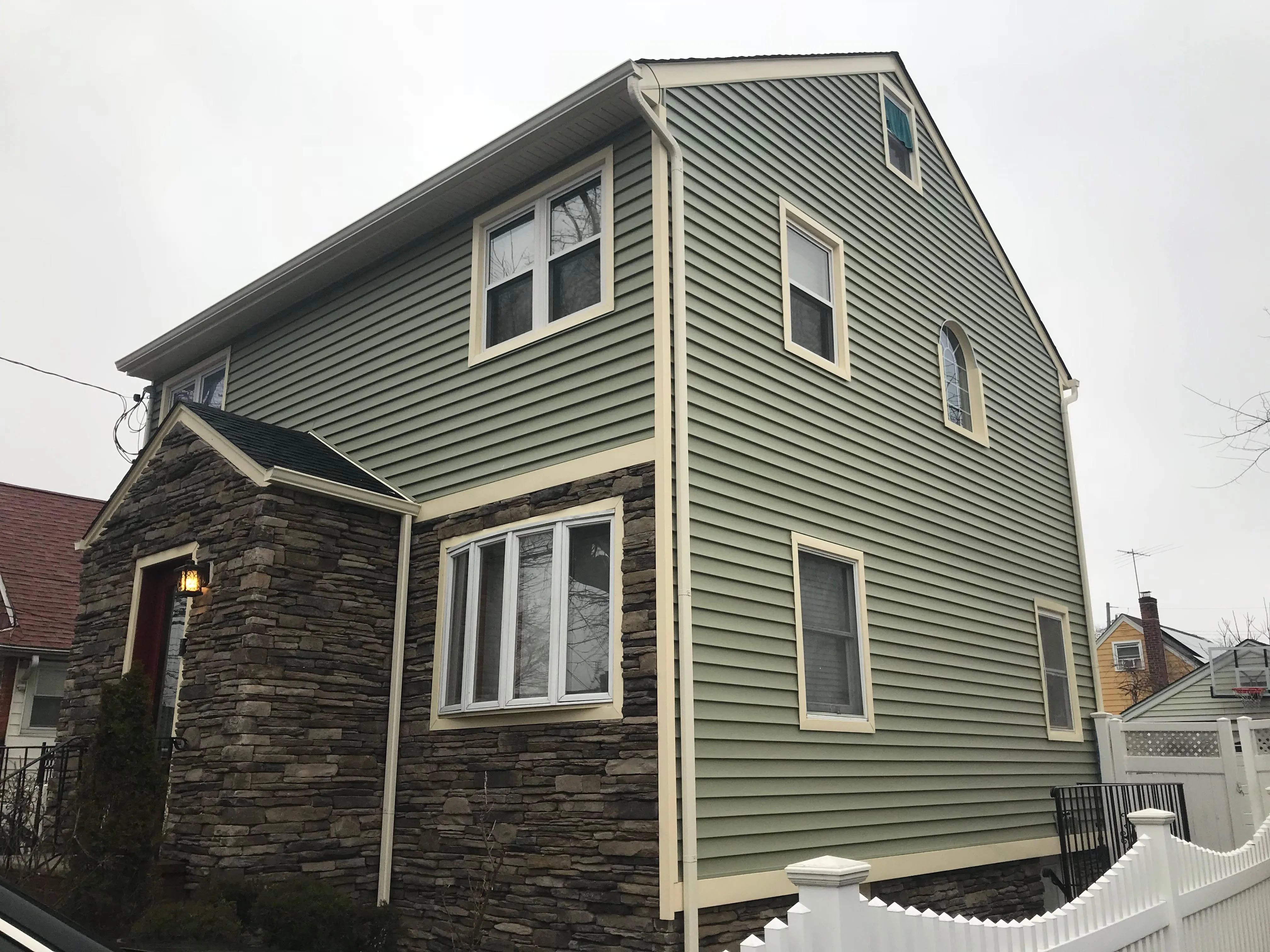Colonial home with sage green vinyl siding, stacked stone veneer accent on front corner, and white vinyl fence