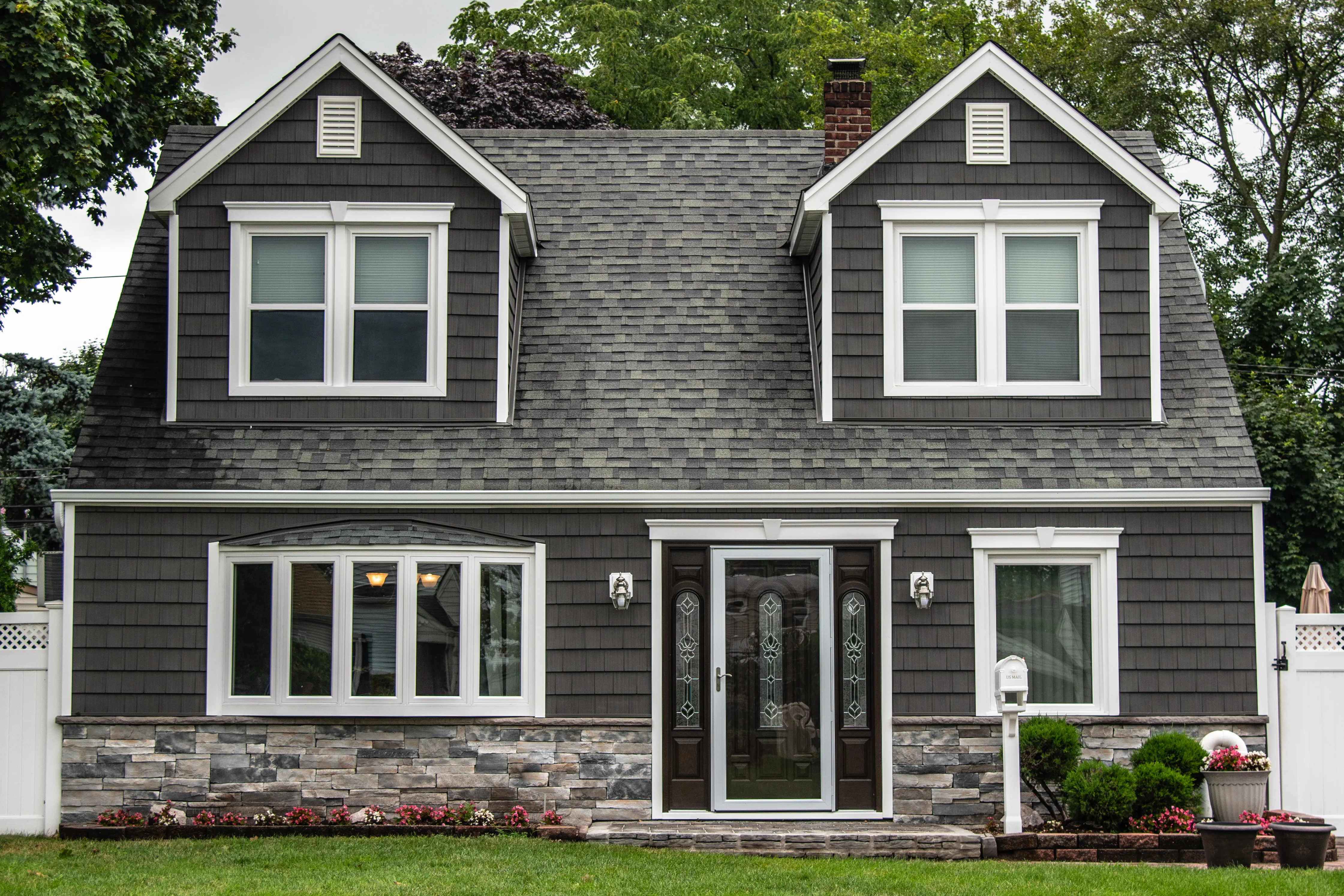 Dutch colonial home with dark gray shake-style siding, white trim, and stone veneer foundation