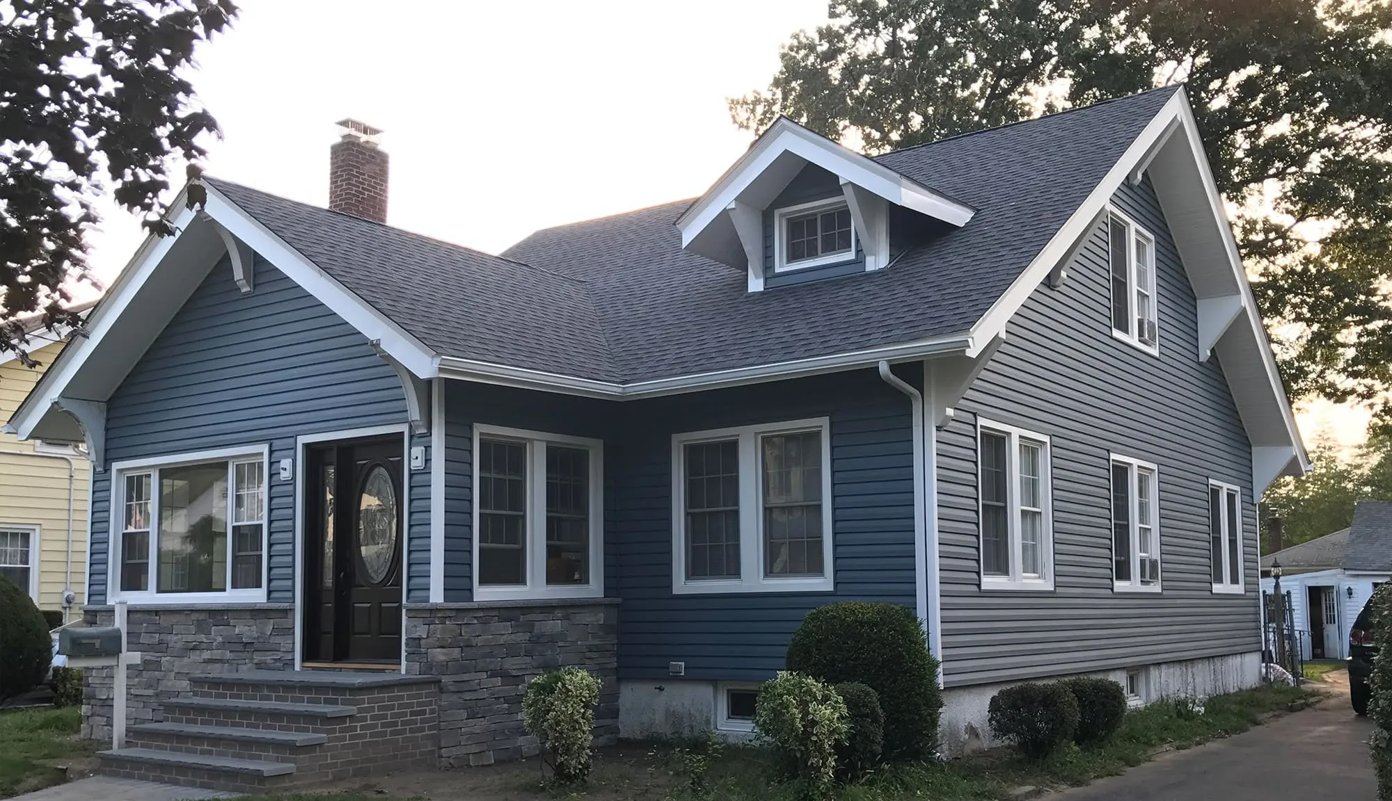 Blue vinyl-sided cape cod with stone veneer foundation, white trim, and decorative oval door glass