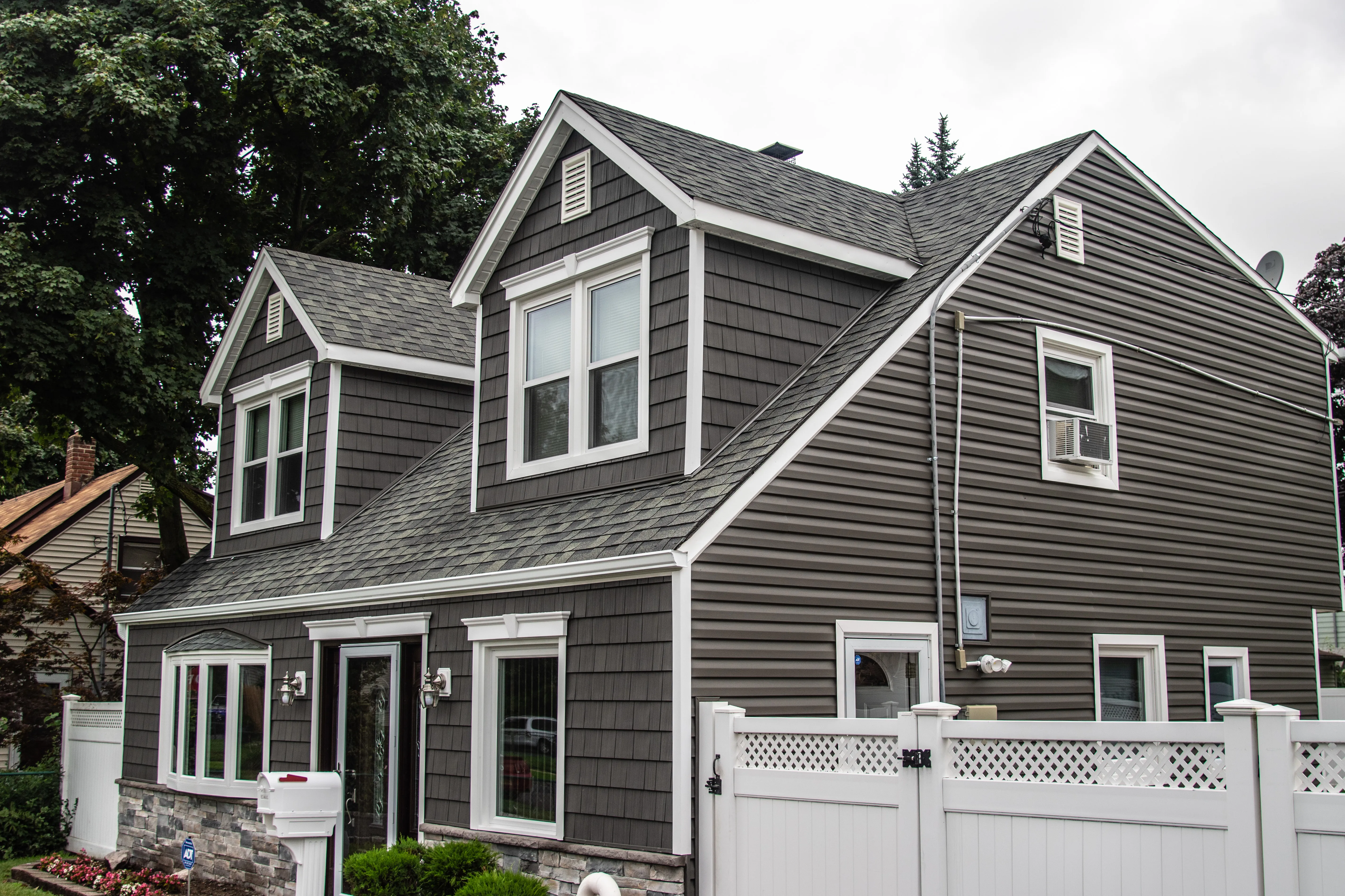 Two-story home with dark charcoal mixed shake and clapboard siding, stone veneer foundation, and white fence
