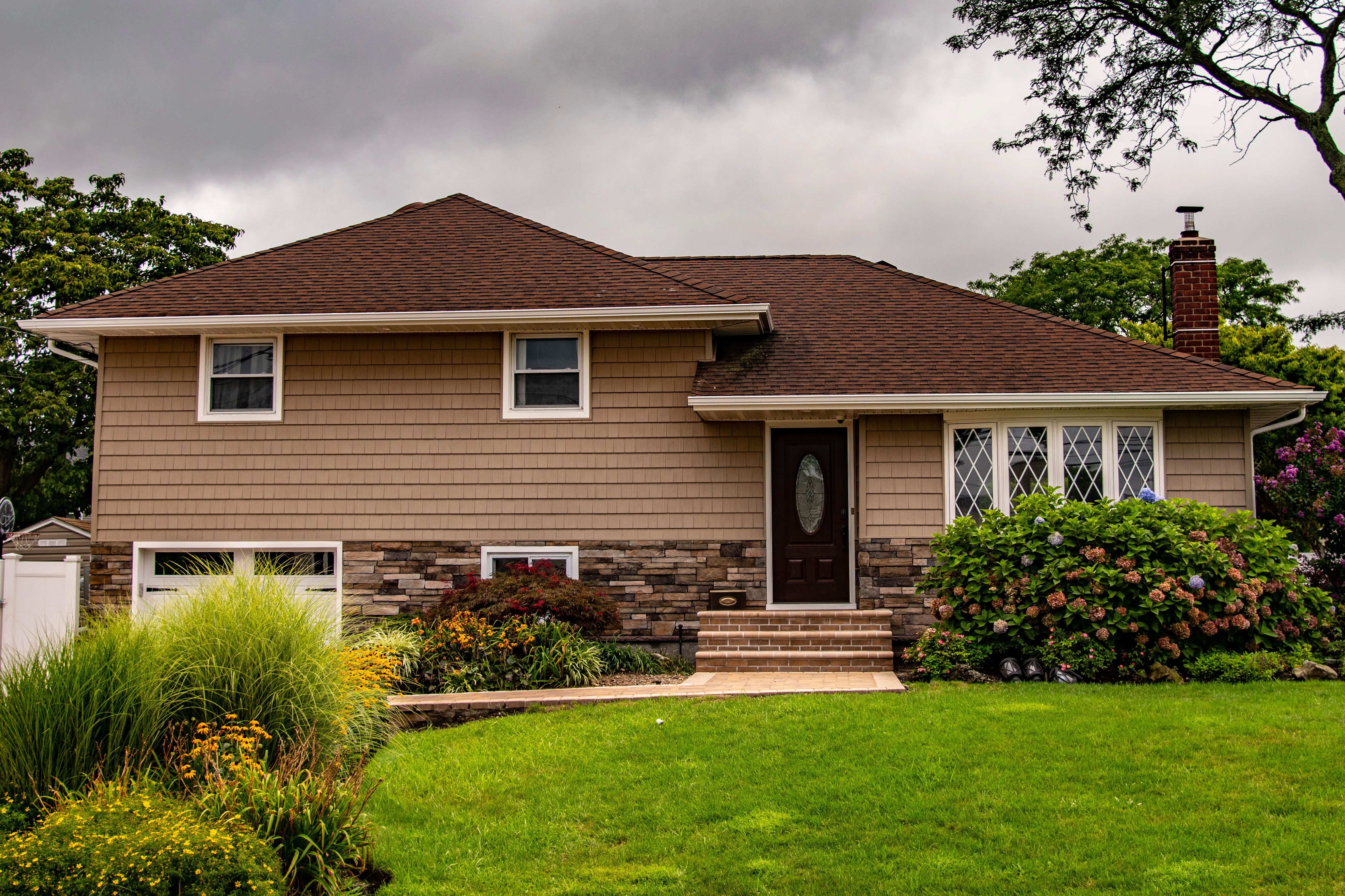 Split-level home with tan shake siding, stone veneer entry, diamond accent windows, and lush landscaping