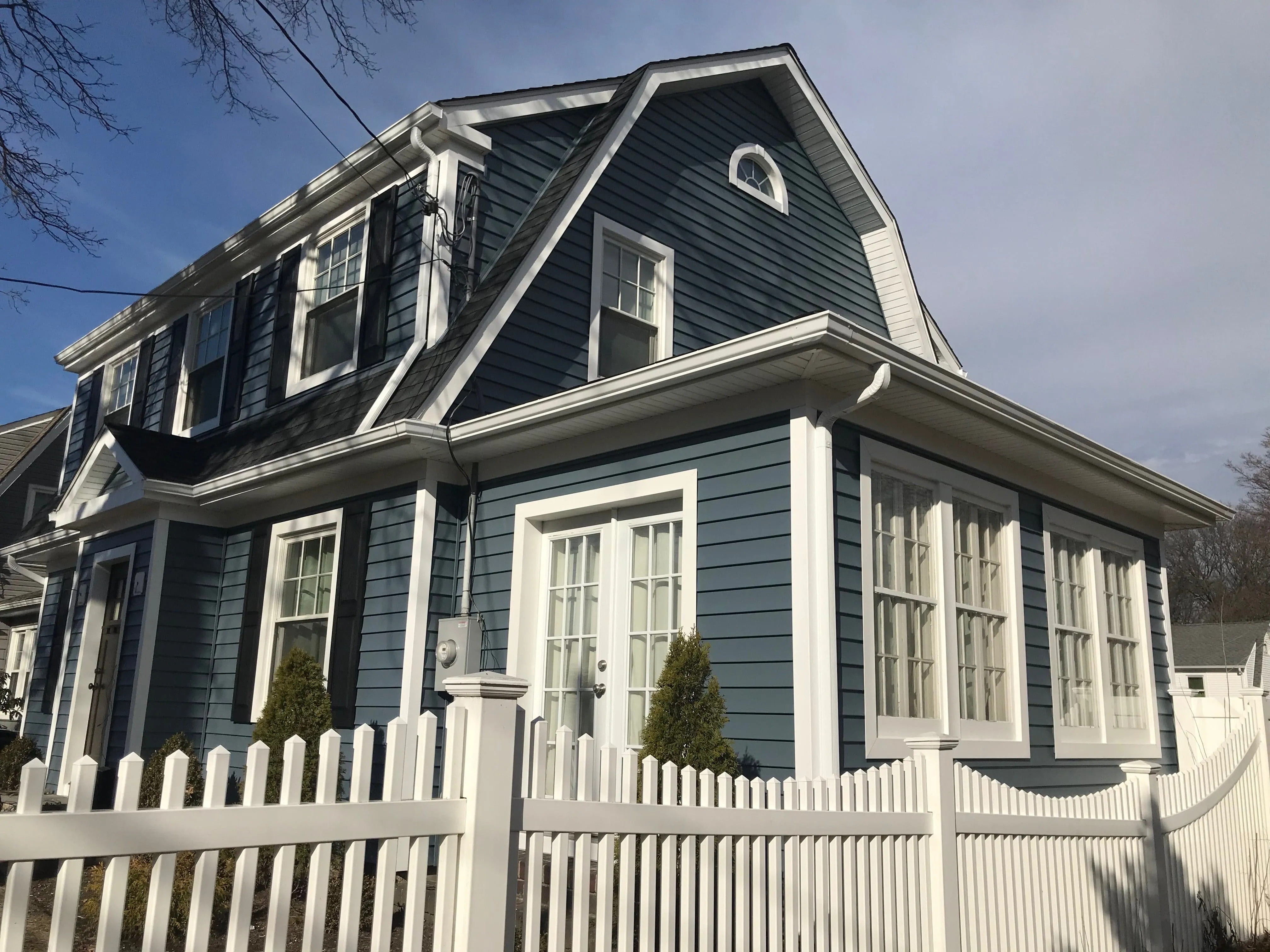 Dutch colonial home with deep blue-green clapboard siding, white trim, and white picket fence